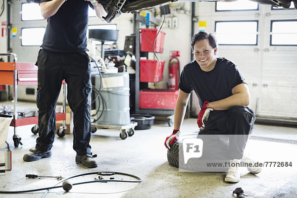 Porträt eines jungen Mechanikers  der auf einem Reifen sitzt  mit einem Mitarbeiter  der das Auto repariert.