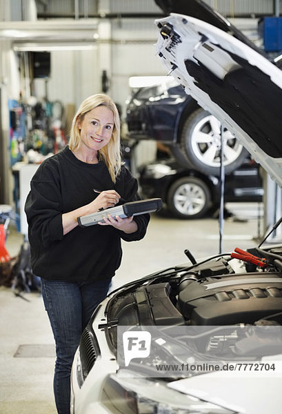 Porträt einer mittleren erwachsenen Mechanikerin  die ein digitales Tablett hält  während sie bei geöffneter Motorhaube steht.