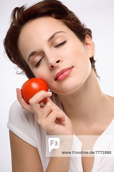 Porträt einer jungen brünetten Frau  Augen geschlossen  Tomate gegen Gesicht haltend
