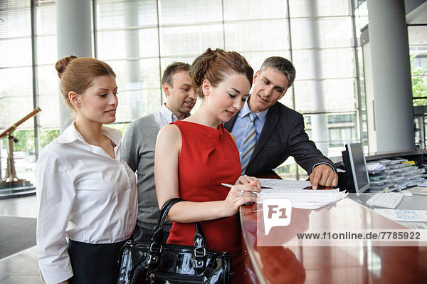Woman signing agreement on reception desk surrounded by group of business people