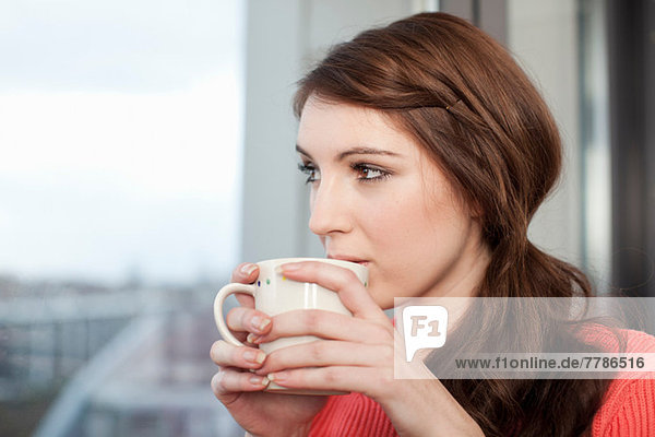 Junge Frau mit Kaffeetasse aus dem Fenster schauend