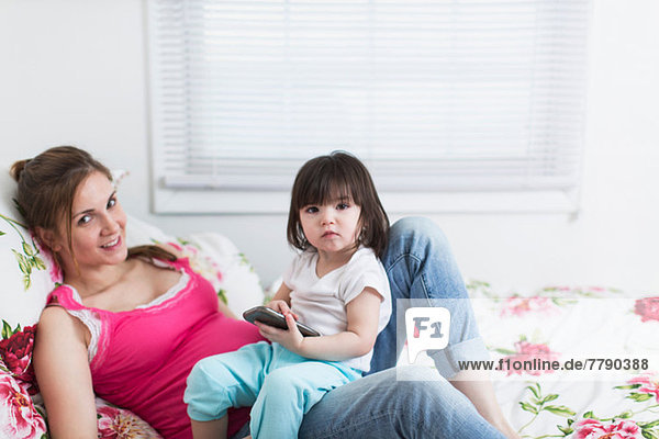 Porträt einer schwangeren Frau und einer Kleinkind-Tochter  die mit einem Smartphone auf dem Bett liegt.
