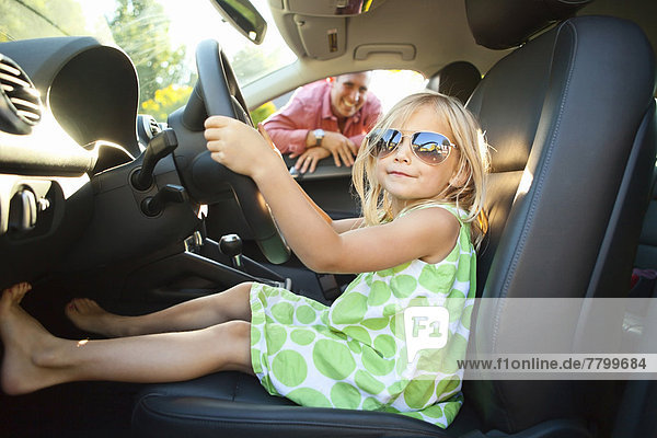 Portrait of little girl sitting in driver's seat of car  pretending to be old enough to drive as her smiling father watches on on a sunny summer evening in Portland  Oregon  USA