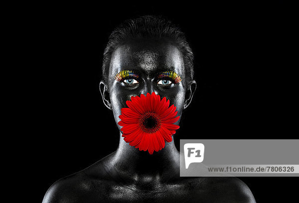 Gesicht einer jungen Frau  bemalt mit schwarzer Farbe  mit bunten Wimpern und einer Blume im Mund