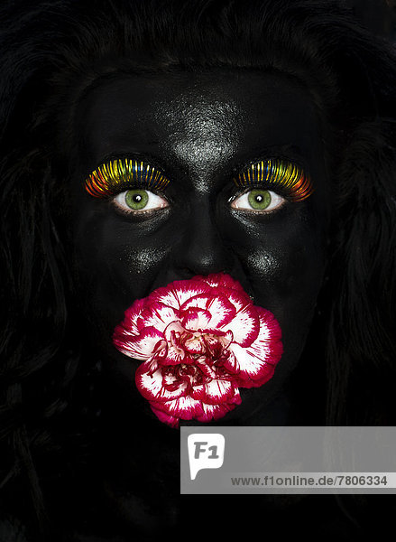 Gesicht einer jungen Frau  bemalt mit schwarzer Farbe  mit bunten Wimpern und einer Blume im Mund