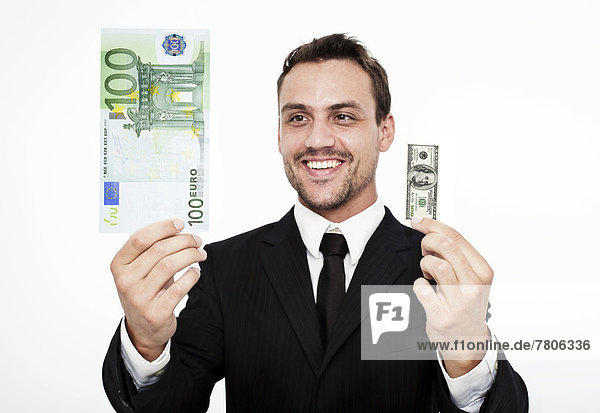 Smiling young man wearing a suit holding a large 100 euro banknote and a small 100 dollar bill