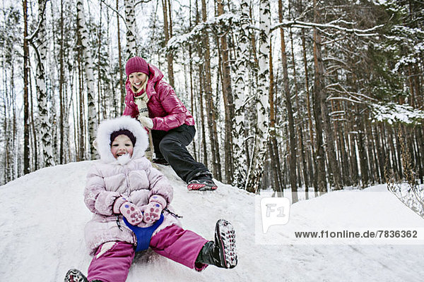 A young girl sledding while her mother looks on