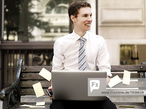 Smiling businessman holding smart phone while using a laptop sitting on bench with sticky notes