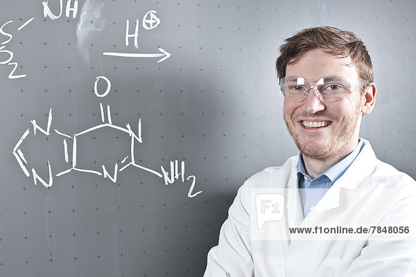 Germany  Portrait of young scientist standing next to chemical equation on chalk board  smiling
