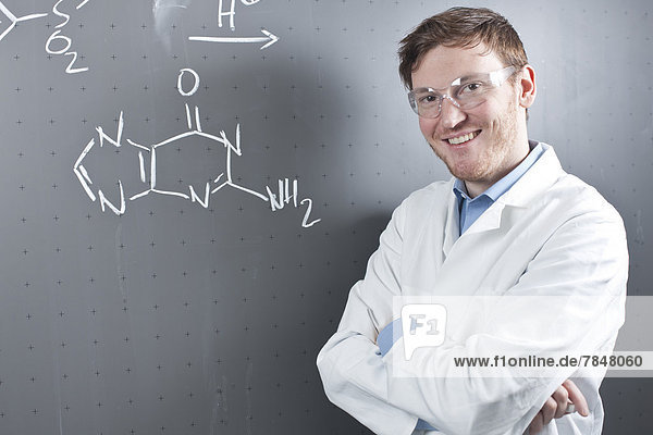 Germany  Portrait of young scientist standing next to chemical equation on chalk board  smiling