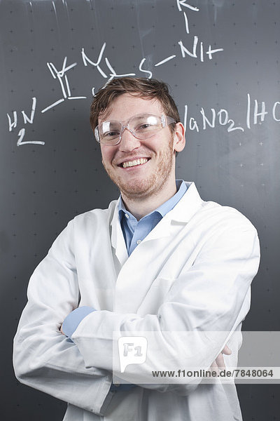 Germany  Portrait of young scientist standing in front of chemical equation on chalk board  smiling