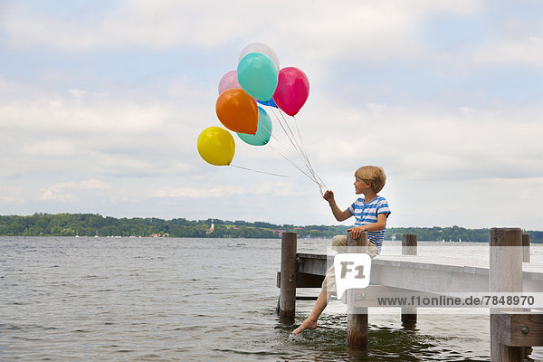 Boy sitting on pier and holding colourful balloons at Starnberg Lake