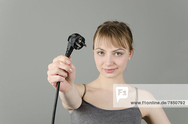 Portrait of young woman holding power plug against grey background