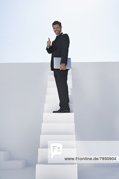 Porträt eines Geschäftsmannes im schwarzen Anzug  der mit Laptop auf der Treppe steht und Daumen nach oben zeigt  lächelnd