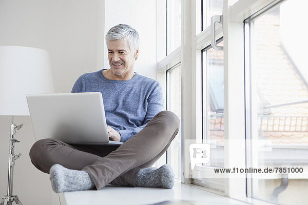 Erwachsener Mann am Fenster sitzend und mit Laptop