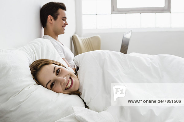 Junge Frau auf dem Bett liegend  junger Mann mit Laptop im Hintergrund