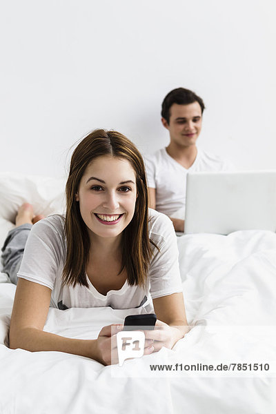 Porträt einer jungen Frau mit Handy  während ein junger Mann mit Laptop im Hintergrund lächelt.