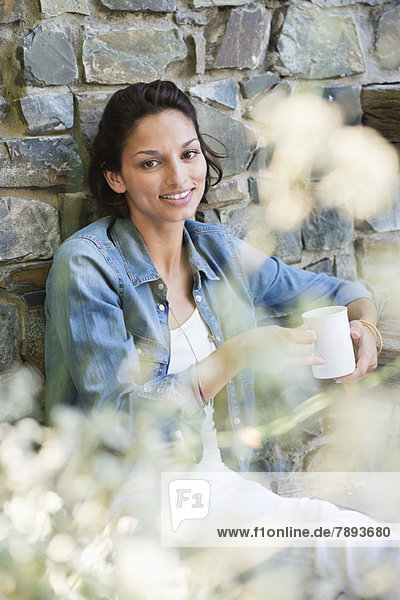 Porträt einer lächelnden Frau  die sich an eine Wand lehnt und Kaffee trinkt.