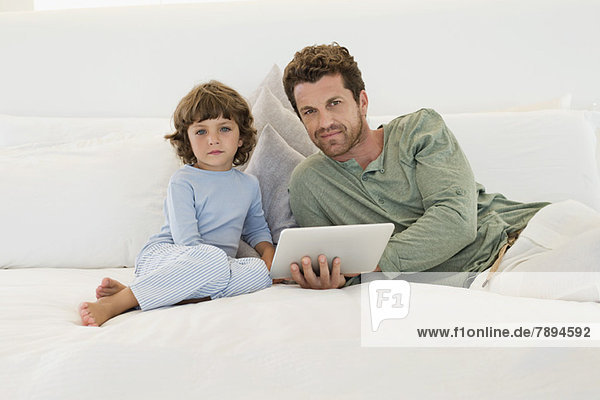 Mann mit einem digitalen Tablett auf dem Bett sitzend mit seinem Sohn