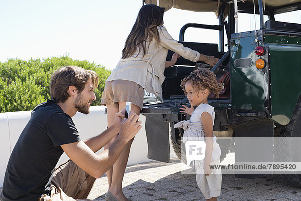 Ein Mann fotografiert seine Tochter mit einem Smartphone neben einem SUV.