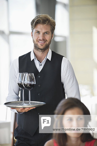 Frau sitzt in einem Restaurant mit einem Kellner  der im Hintergrund ein Tablett mit Weingläsern hält.