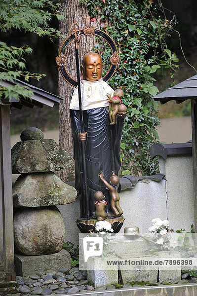 Buddhist Jizo statue with bib and halo  the patron god of the children  with a child in his arms and children at his feet  next to a stone lamp