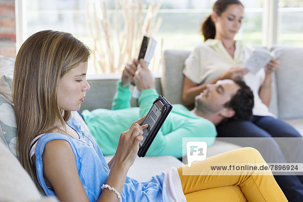 Mädchen,  das ein digitales Tablett benutzt,  während ihre Eltern zu Hause Bücher lesen.