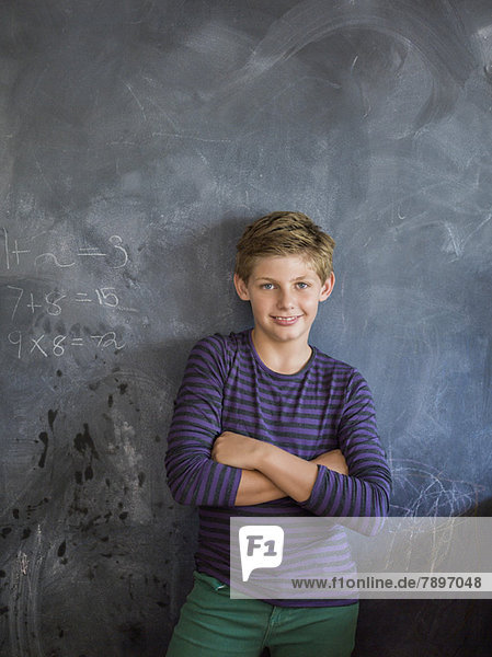Junge lächelt mit verschränkten Armen vor einer Tafel im Klassenzimmer.