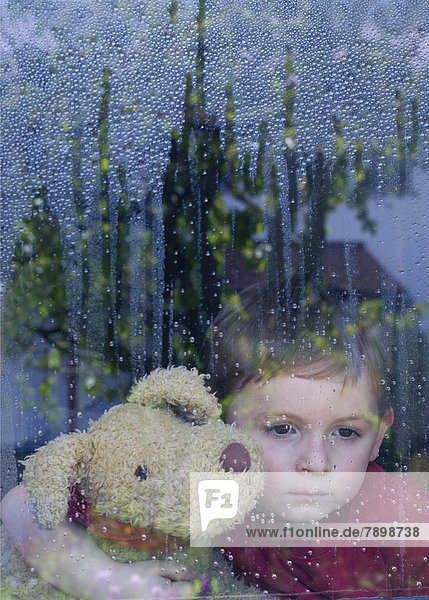 Junge schaut traurig aus Fensterscheibe mit Wassertropfen Kuscheltier im Arm