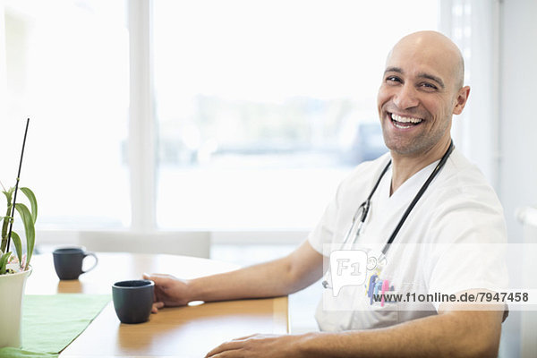 Portrait of happy male doctor with coffee cup sitting at desk in hospital
