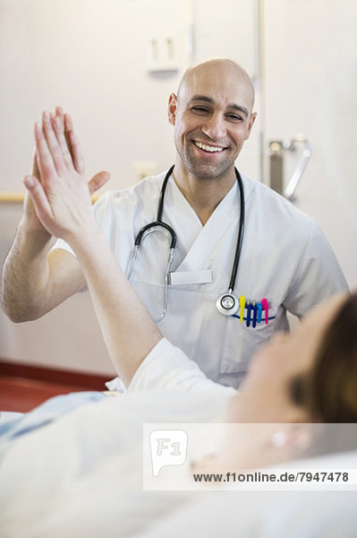Mid adult male doctor giving high five to female patient lying on bed in hospital ward