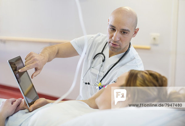Mittlerer erwachsener männlicher Arzt  der dem auf dem Bett liegenden Patienten auf der Krankenstation eine digitale Tablette zeigt.