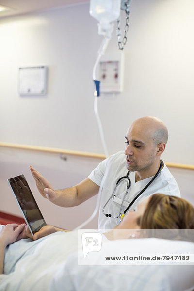 Mittlerer erwachsener männlicher Arzt,  der dem auf dem Bett liegenden Patienten auf der Krankenstation über eine digitale Tablette erklärt.