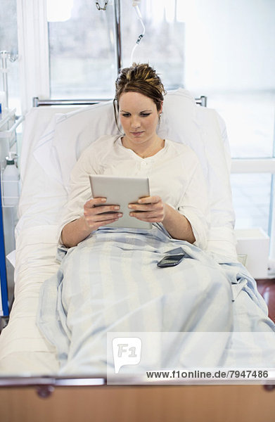 Mittlere erwachsene Patientin  die eine Tablette benutzt  während sie sich auf dem Bett in der Krankenstation entspannt.