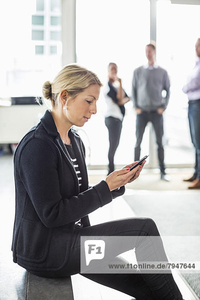 Seitenansicht der mittleren erwachsenen Geschäftsfrau per Handy mit Kollegen im Hintergrund im Büro