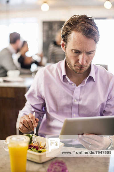 Geschäftsmann  der beim Frühstück mit Kollegen im Hintergrund im Restaurant das digitale Tablett betrachtet