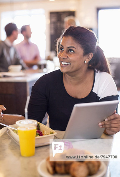 Geschäftsfrau schaut weg  während sie das digitale Tablett am Restauranttisch hält  mit einem Kollegen im Hintergrund.
