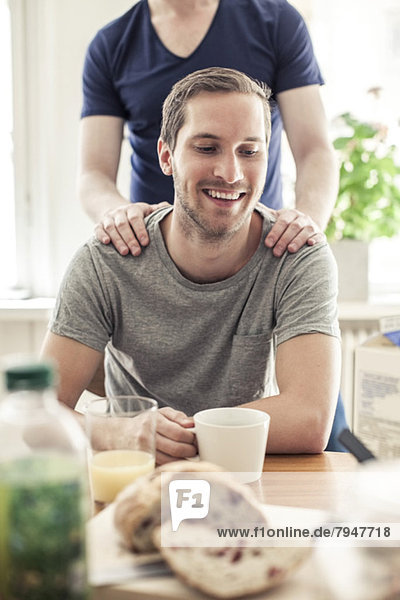 Fröhlicher schwuler Mann beim Kaffee  während der Partner hinter ihm am Tisch zu Hause steht.