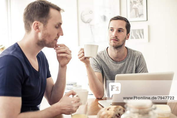 Homosexual couple having breakfast together with laptop at table in home