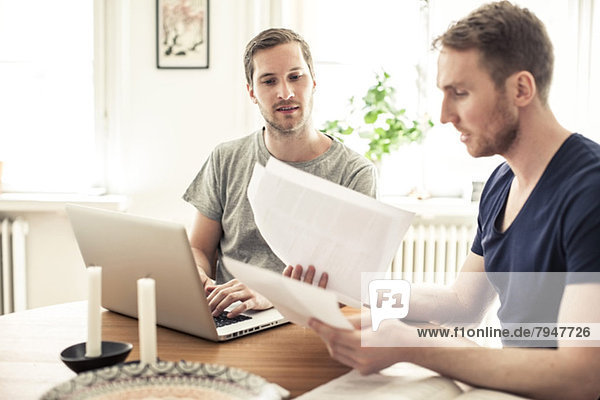 Gay man reading paper while partner using laptop at table in home