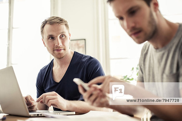 Young gay man looking at partner using mobile phone at table in house