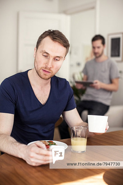 Junger schwuler Mann mit Handy beim Frühstück am Tisch mit Partner im Hintergrund