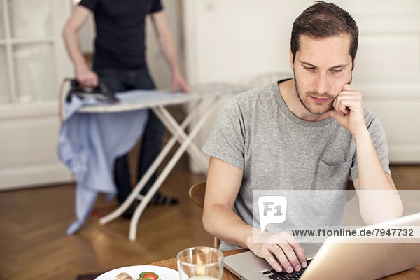 Homosexuell Mann mit Laptop am Tisch mit Partner Bügeln im Hintergrund