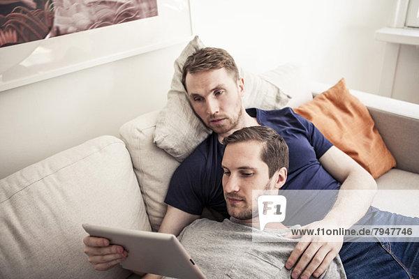 Junge Homosexuelle nutzen gemeinsam digitale Tabletten und entspannen sich zu Hause auf dem Sofa.