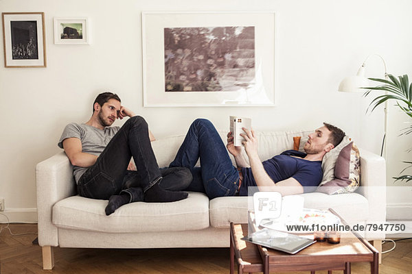 Young gay man reading book while partner looking at him on sofa