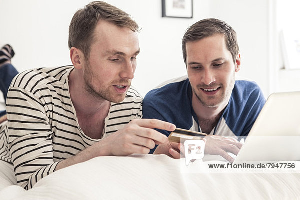 Homosexuelles Paar  das zusammen auf dem Laptop online einkauft  während es zu Hause im Bett liegt.