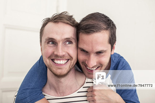 Fröhlicher junger schwuler Mann schaut weg  während der Partner zu Hause von hinten umarmt wird.
