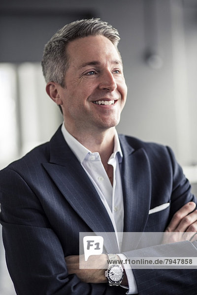 Thoughtful mature businessman smiling while standing arms crossed in office