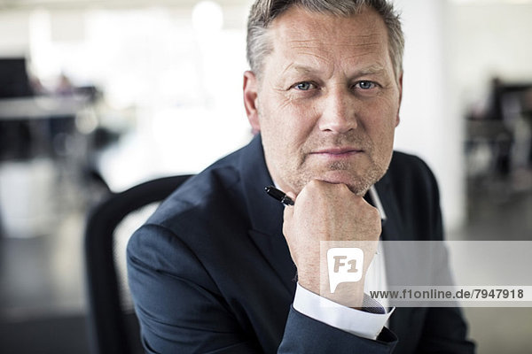 Portrait of confident mature businessman sitting with hand on chin at office