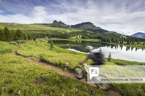 A mountain biker rides by in a blur through green mountain scenery with a lake in the background.